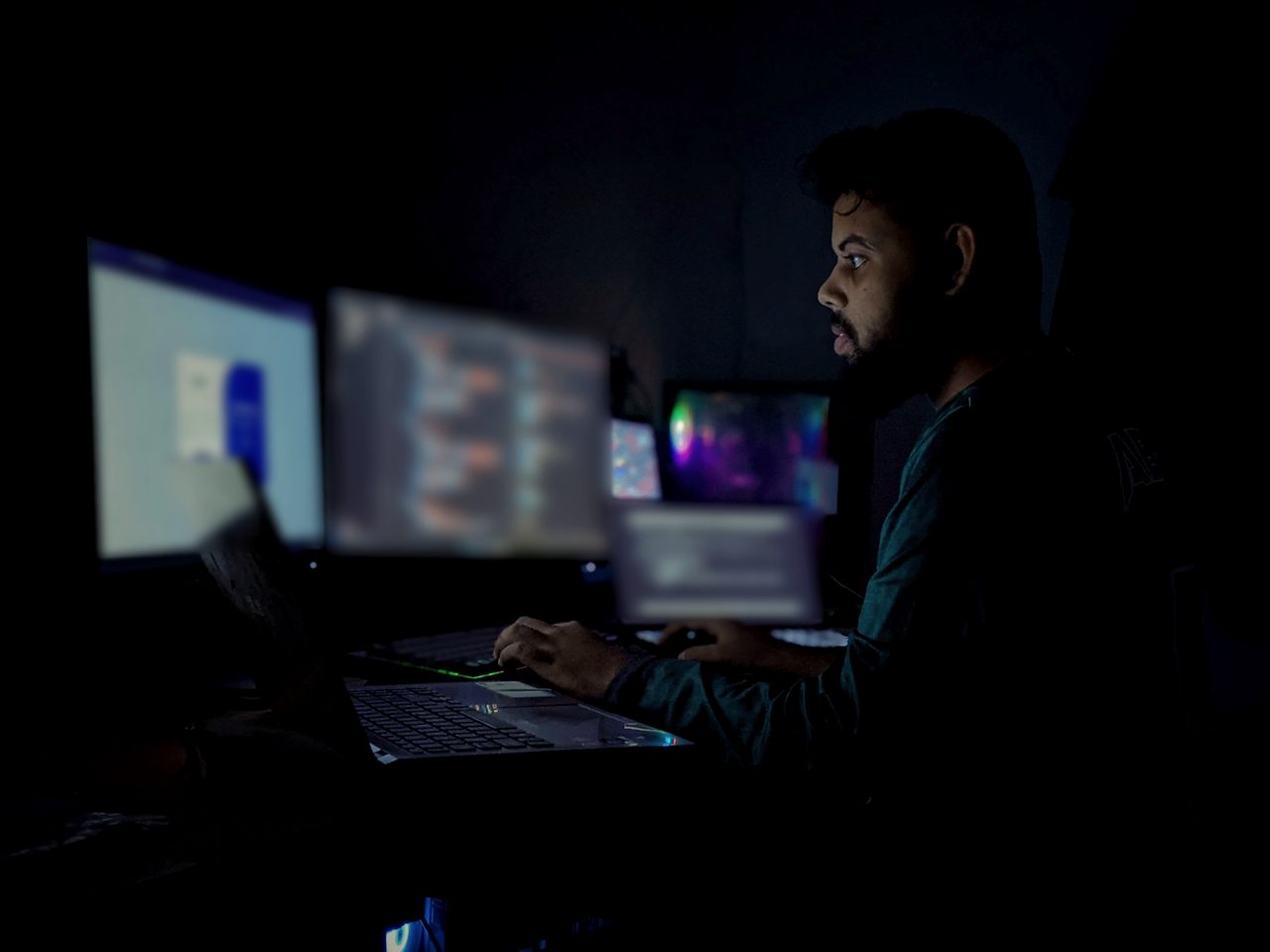 A bearded man in profile works at a desk with three monitors and a laptop in a dark room, lit by the screens.