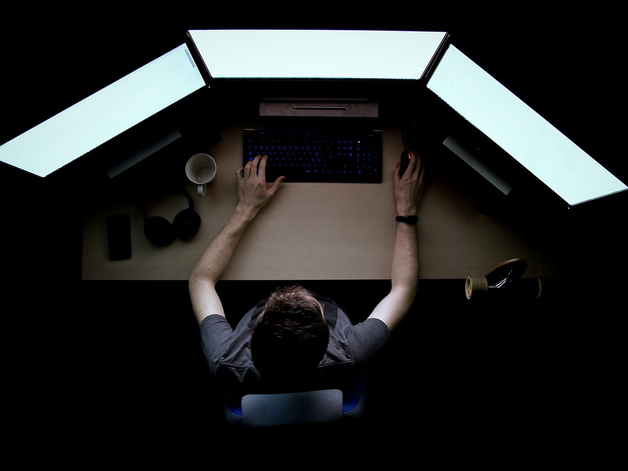 Top-down view of a man at a desk surrounded by three glowing monitors, hands on a backlit keyboard and a mouse.