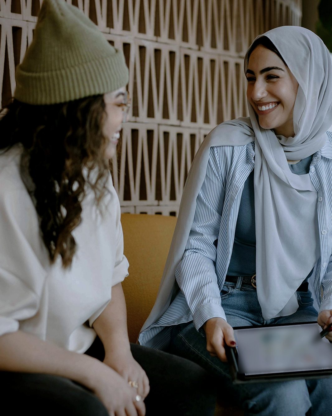 شابتان تتحدثان وتضحكان على أريكة؛ إحداهما بحجاب فاتح تحمل جهازًا لوحيًا وقلمًا رقميًا.
