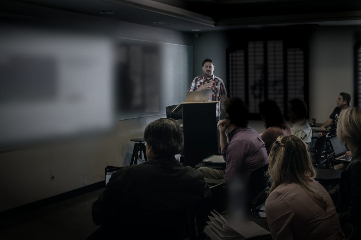 In a dark training room, a facilitator at a lectern beside a projected screen talks to a small group seated in the foreground.