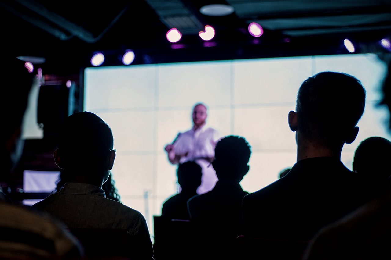 From the back of a dark room, seated audience members are silhouetted against a large bright screen showing a speaker.