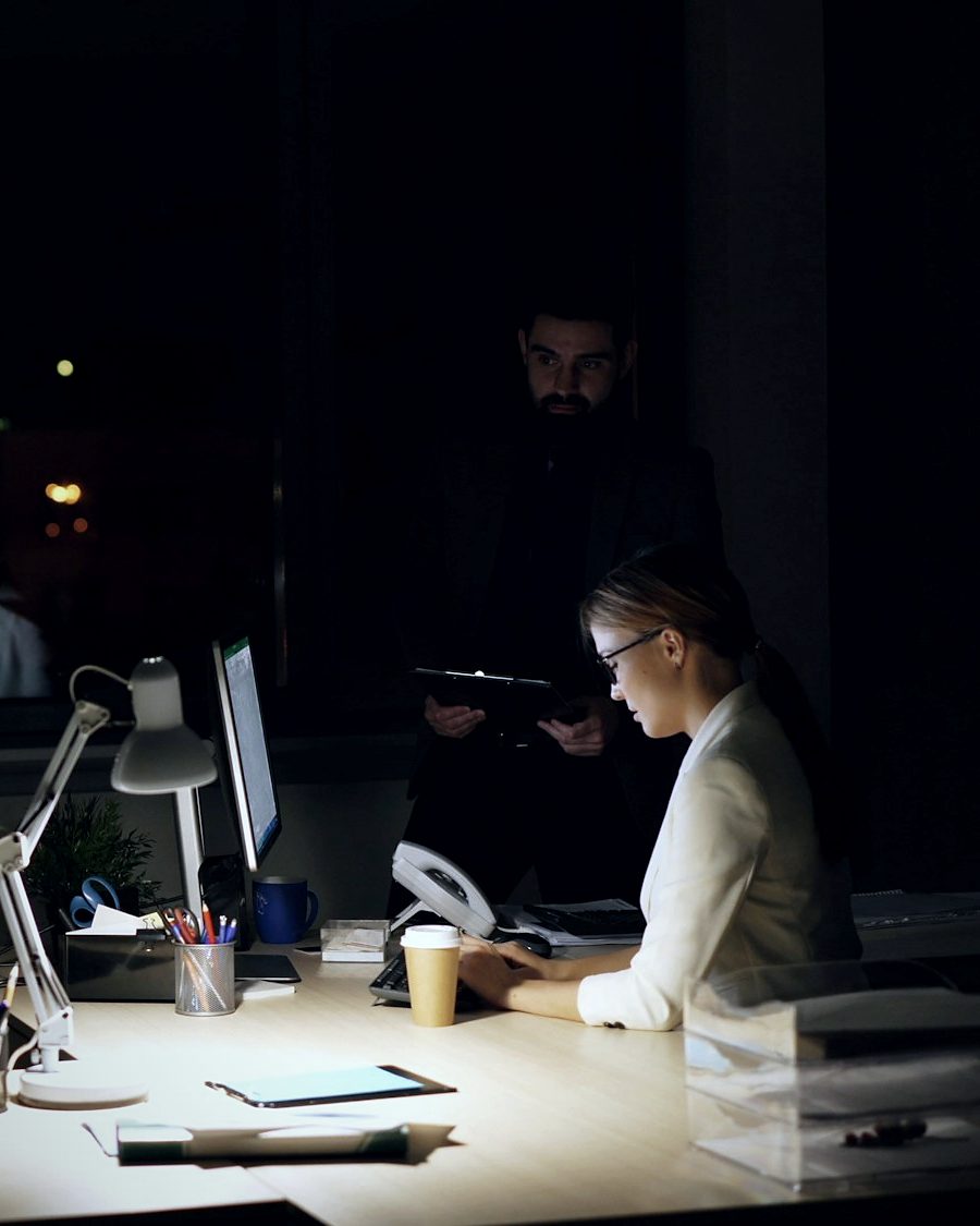 In a dark office at night, a woman types at a lamp-lit desk while a man stands behind her holding a tablet.